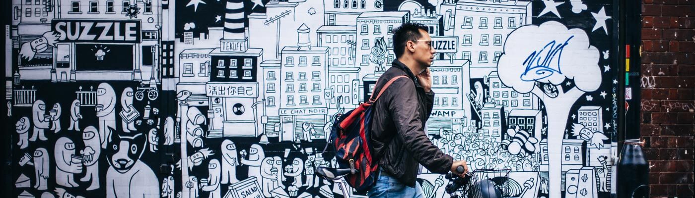 A man with his bike and his phone on his ear walks past a wall with street art 